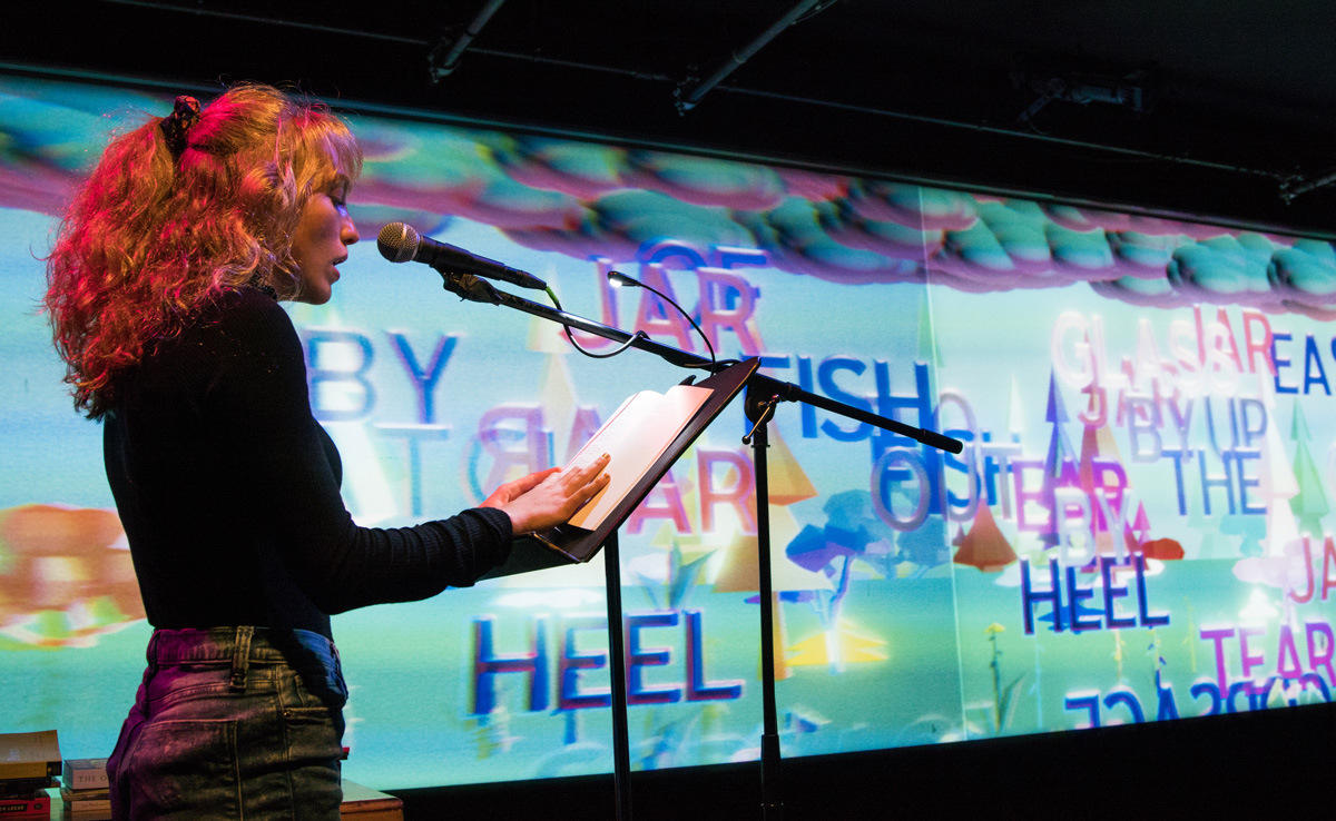 Poet Rhiannon McGavin reading in front of a neon landscape projection